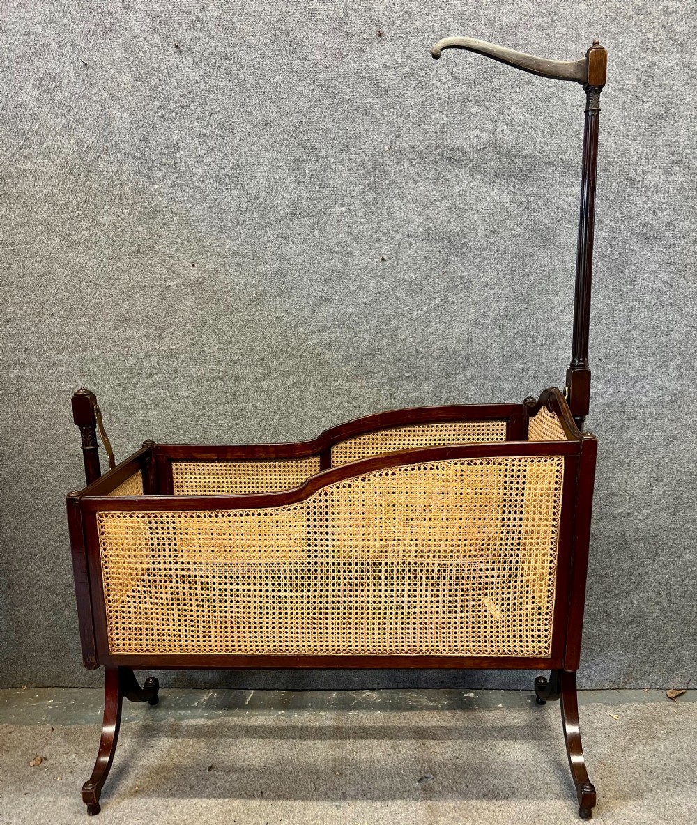 An antique Edwardian dark mahogany swinging crib with woven cane side panels and a lace drape suspended from a tall wooden pole. View II