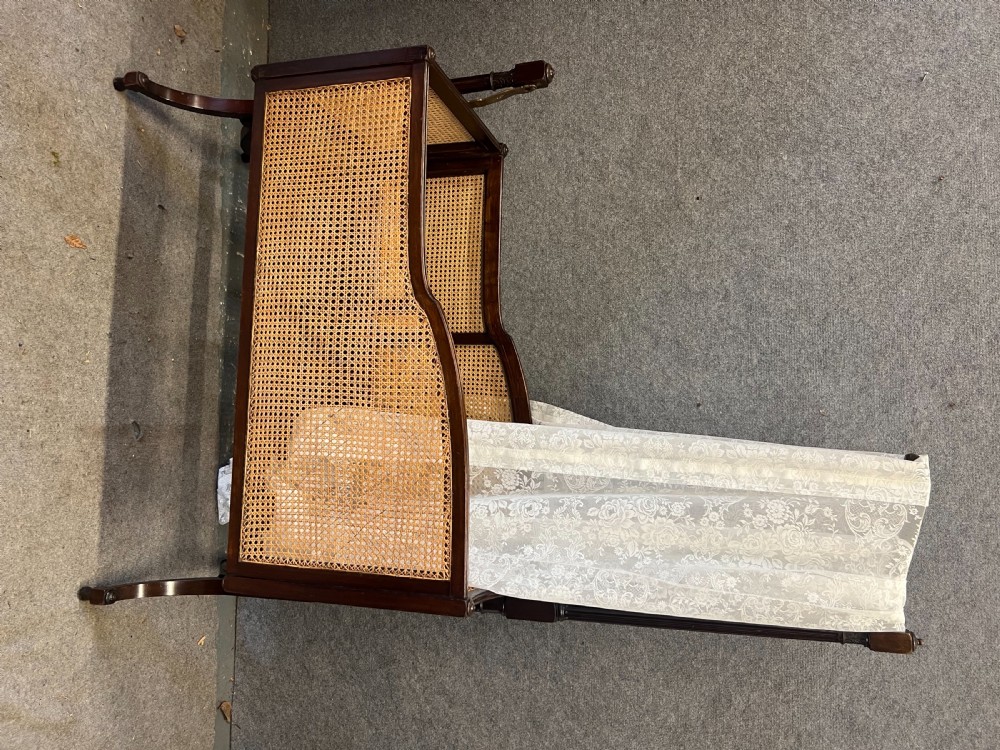 An antique Edwardian dark mahogany swinging crib with woven cane side panels and a lace drape suspended from a tall wooden pole. View III