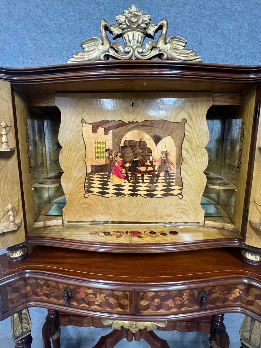 A two-tier dark walnut cocktail cabinet featuring intricate floral marquetry, a serpentine front, gilded carved accents, and turned legs with a contoured stretcher. View VI