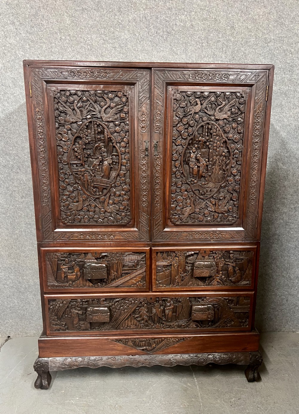 Intricately relief-carved dark camphor wood cabinet-on-chest featuring two upper cupboard doors decorated with figurative scenes above two short and one long drawer.