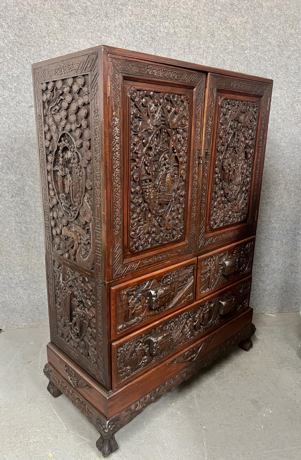 Intricately relief-carved dark camphor wood cabinet-on-chest featuring two upper cupboard doors decorated with figurative scenes above two short and one long drawer. View II