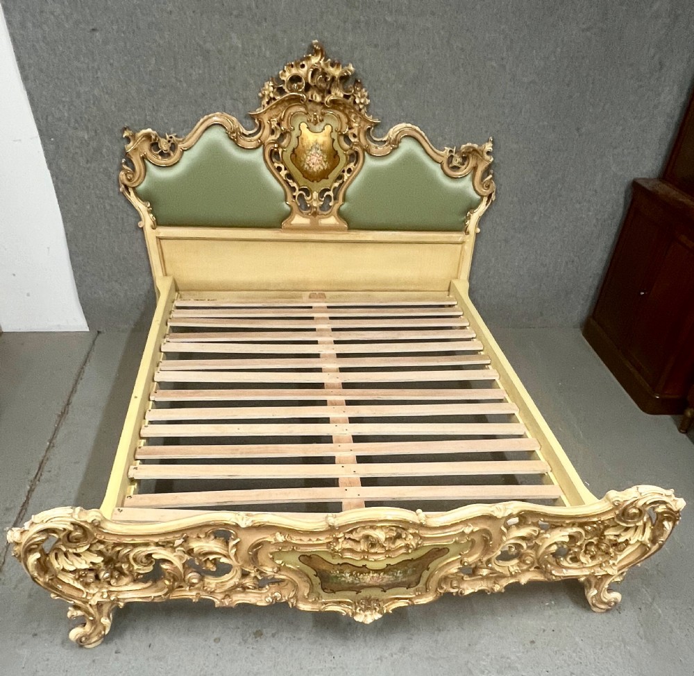 An ornate French Rococo style king size bed frame featuring cream painted and carved wood with gilded accents, sage green upholstered headboard panels, and hand-painted floral cartouches. View IV