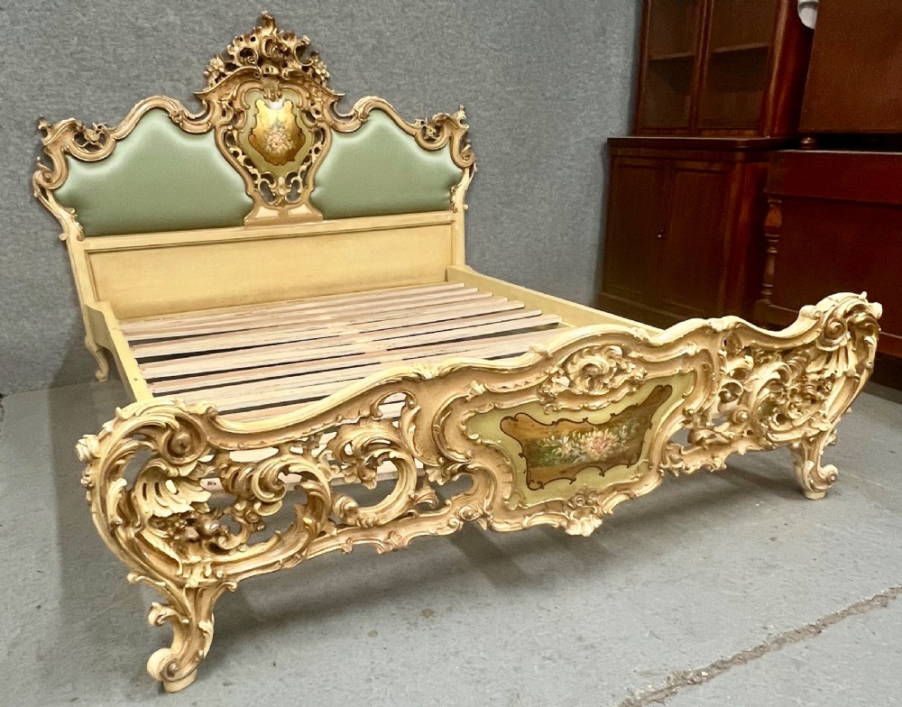 An ornate French Rococo style king size bed frame featuring cream painted and carved wood with gilded accents, sage green upholstered headboard panels, and hand-painted floral cartouches.