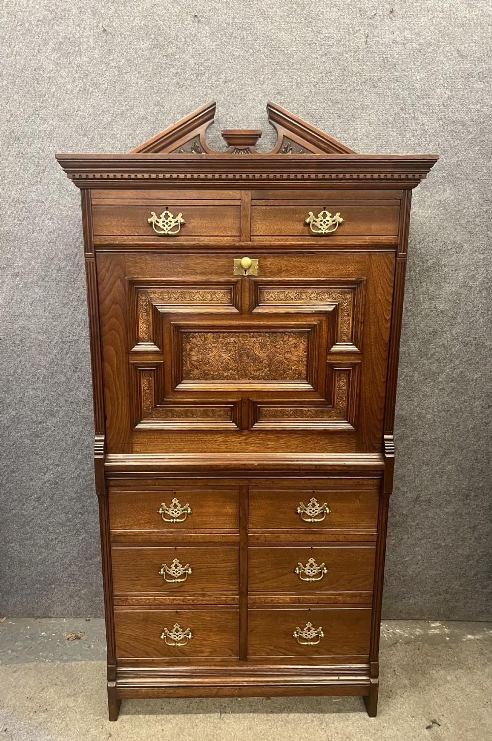 A tall dark timber escritoire desk featuring a fall-front writing surface with geometric panelling, brass drop handles, and multiple storage drawers below. View III