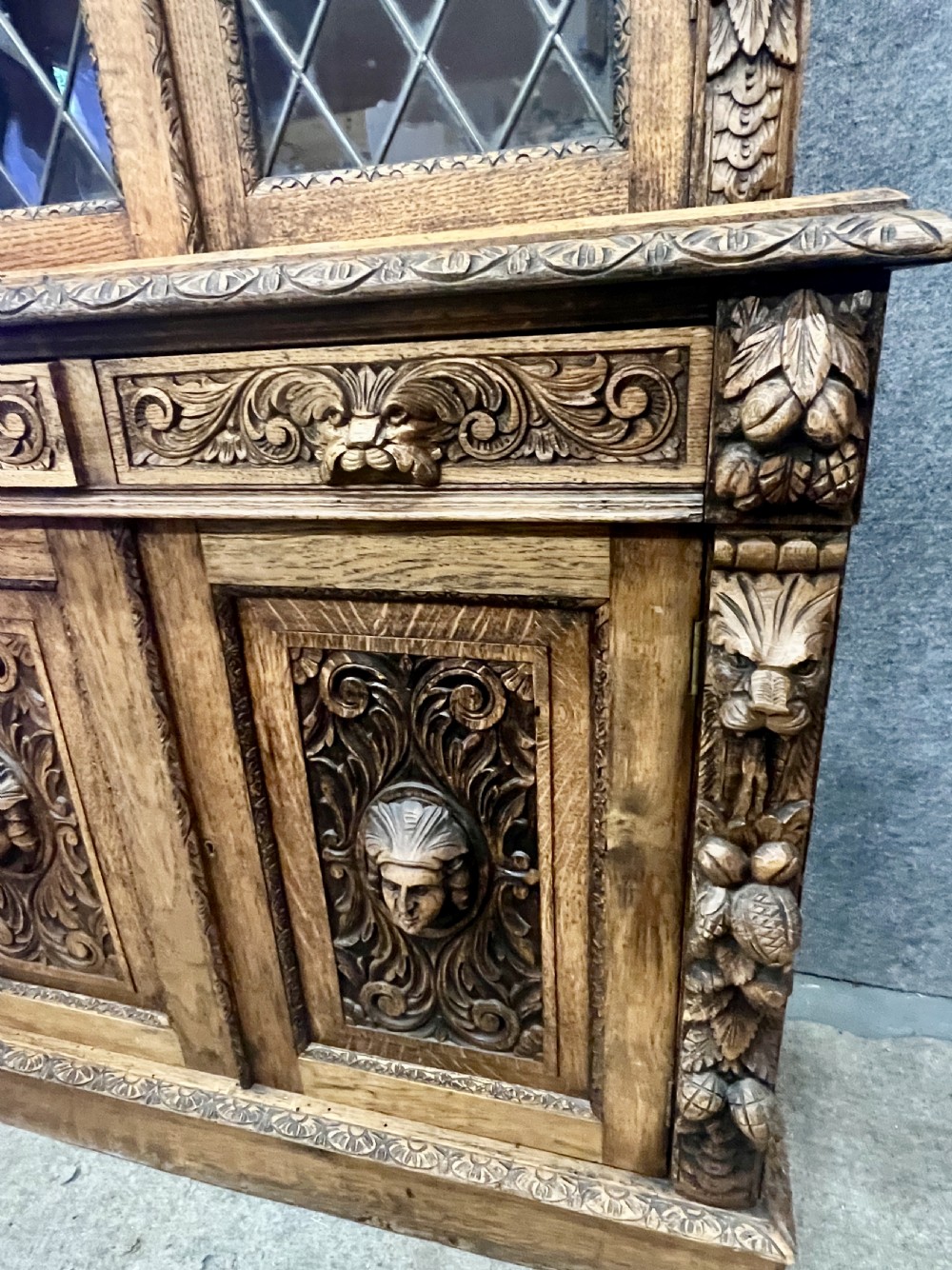 A 19th-century carved oak bookcase featuring diamond leaded glass upper doors, carved figurative figures, two small drawers, and panelled lower doors. View IV