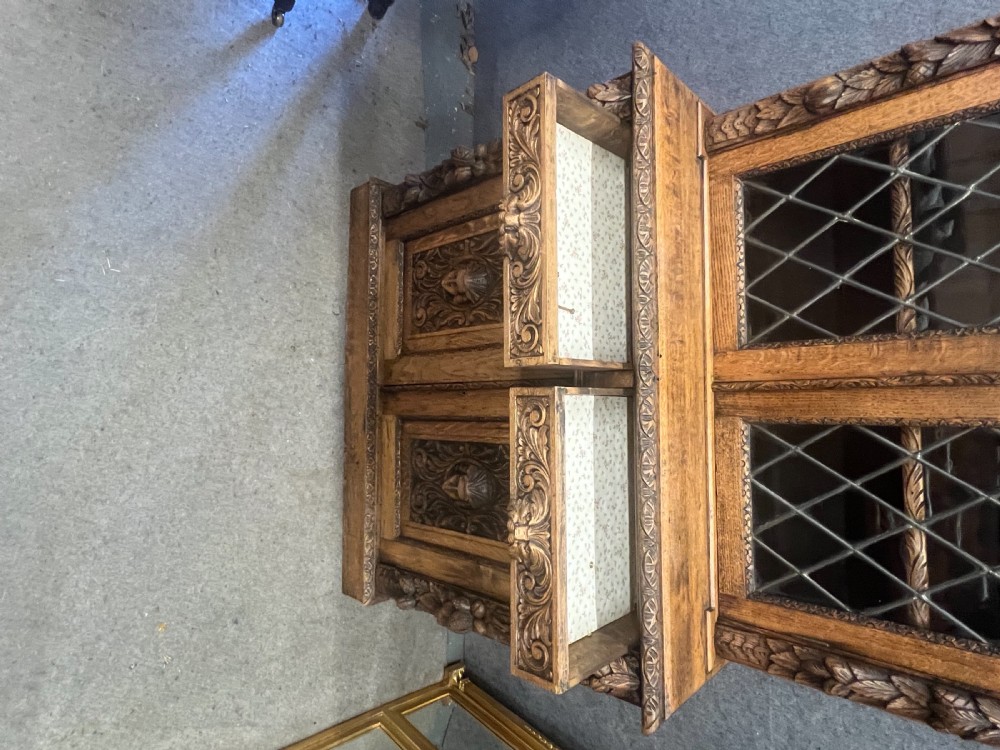 A 19th-century carved oak bookcase featuring diamond leaded glass upper doors, carved figurative figures, two small drawers, and panelled lower doors. View V