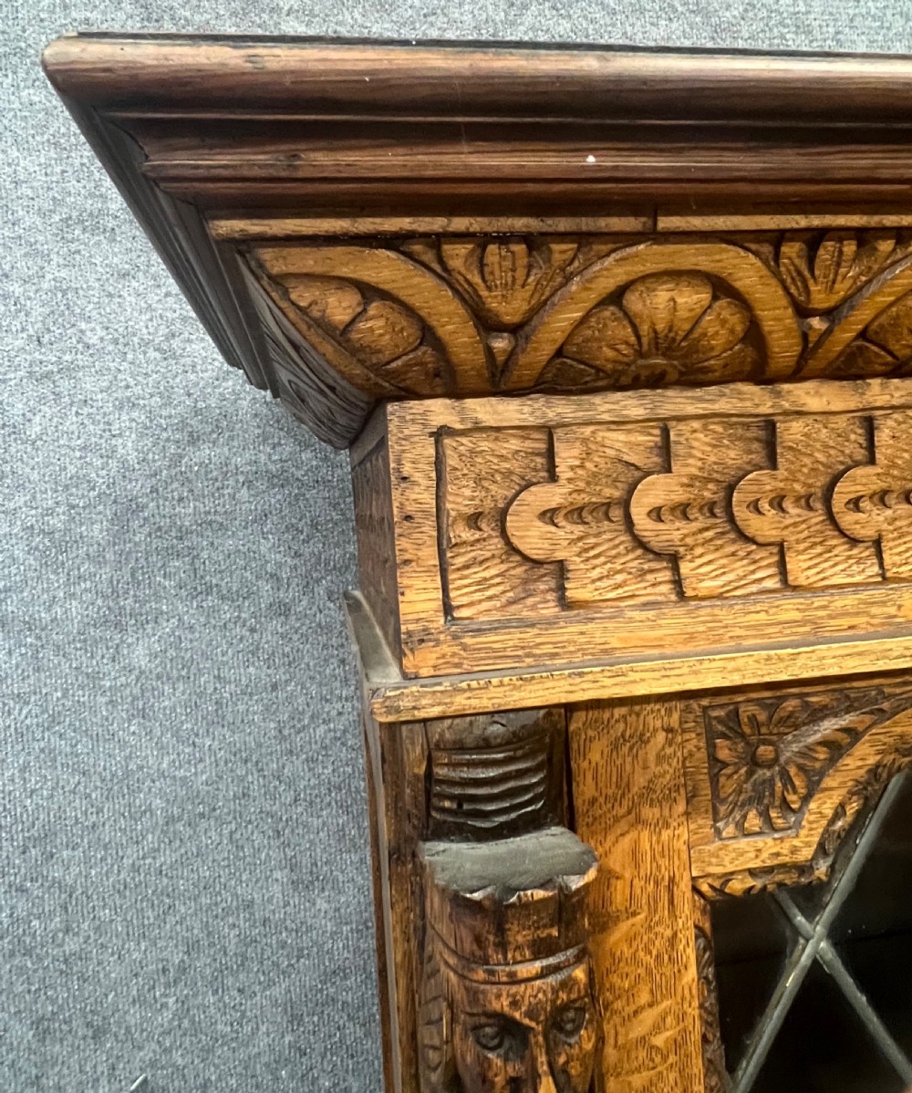 A 19th-century carved oak bookcase featuring diamond leaded glass upper doors, carved figurative figures, two small drawers, and panelled lower doors. View VI