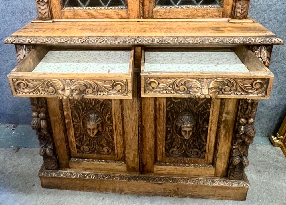 A 19th-century carved oak bookcase featuring diamond leaded glass upper doors, carved figurative figures, two small drawers, and panelled lower doors. View IX