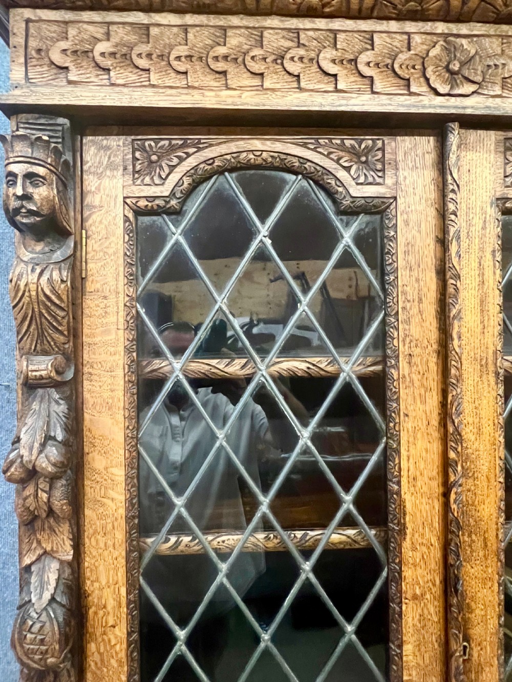 A 19th-century carved oak bookcase featuring diamond leaded glass upper doors, carved figurative figures, two small drawers, and panelled lower doors. View XV