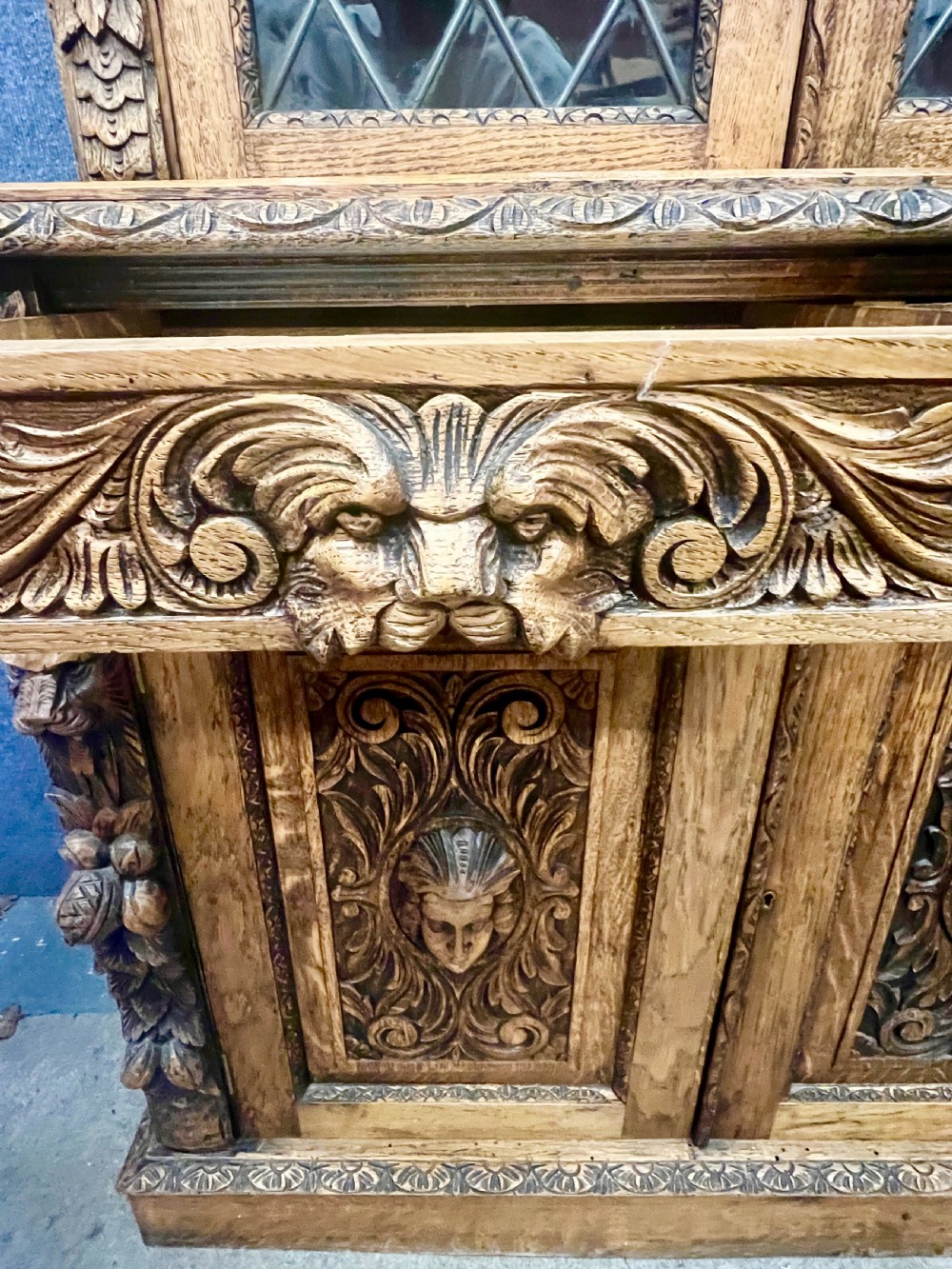 A 19th-century carved oak bookcase featuring diamond leaded glass upper doors, carved figurative figures, two small drawers, and panelled lower doors. View XVI