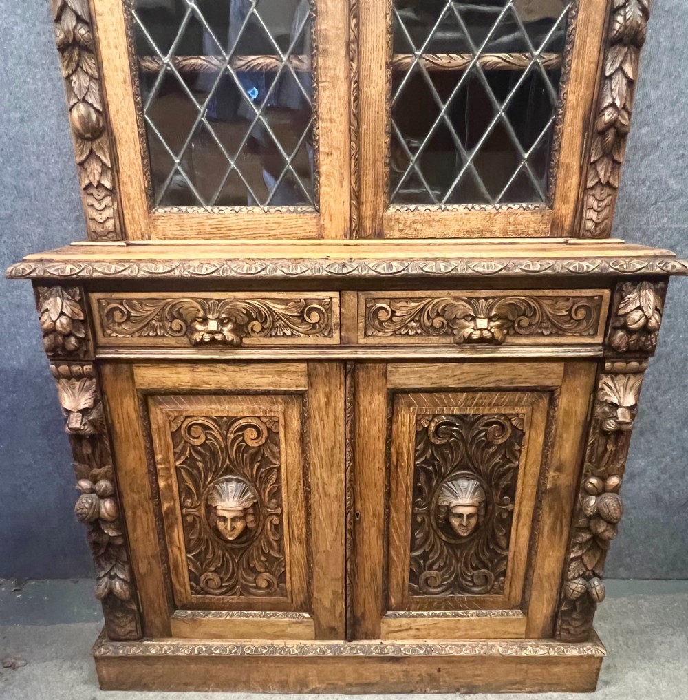 A 19th-century carved oak bookcase featuring diamond leaded glass upper doors, carved figurative figures, two small drawers, and panelled lower doors. View XVII