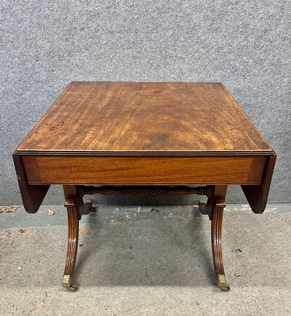 An antique Regency mahogany sofa table featuring two hinged drop leaves, a reeded timber border, twin reeded sabre legs joined by a turned stretcher, and brass caps on castors.