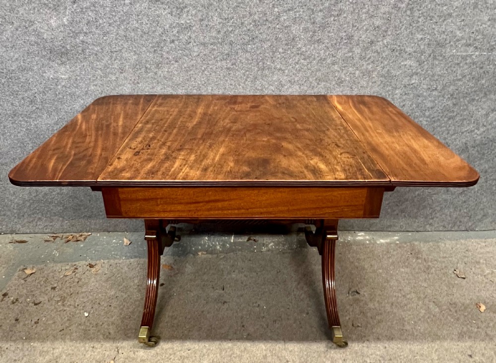 An antique Regency mahogany sofa table featuring two hinged drop leaves, a reeded timber border, twin reeded sabre legs joined by a turned stretcher, and brass caps on castors. View II