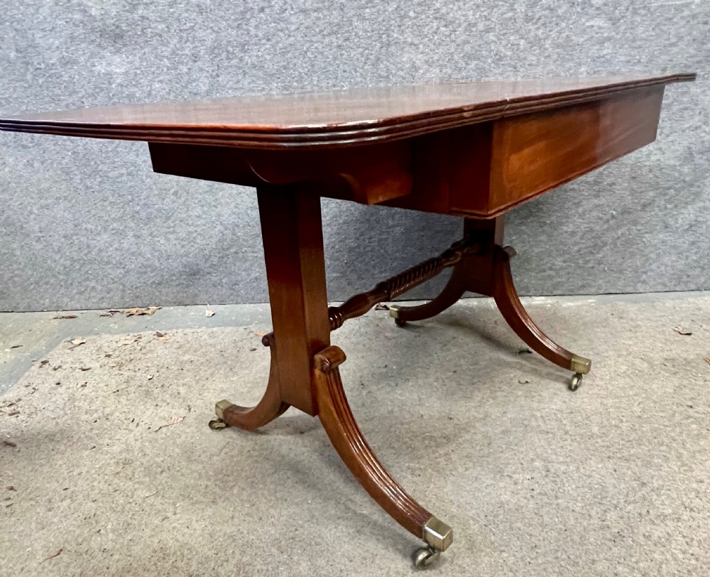 An antique Regency mahogany sofa table featuring two hinged drop leaves, a reeded timber border, twin reeded sabre legs joined by a turned stretcher, and brass caps on castors. View V