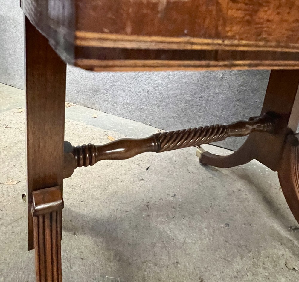 An antique Regency mahogany sofa table featuring two hinged drop leaves, a reeded timber border, twin reeded sabre legs joined by a turned stretcher, and brass caps on castors. View VII
