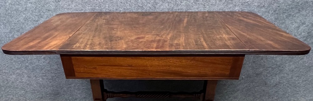 An antique Regency mahogany sofa table featuring two hinged drop leaves, a reeded timber border, twin reeded sabre legs joined by a turned stretcher, and brass caps on castors. View IX