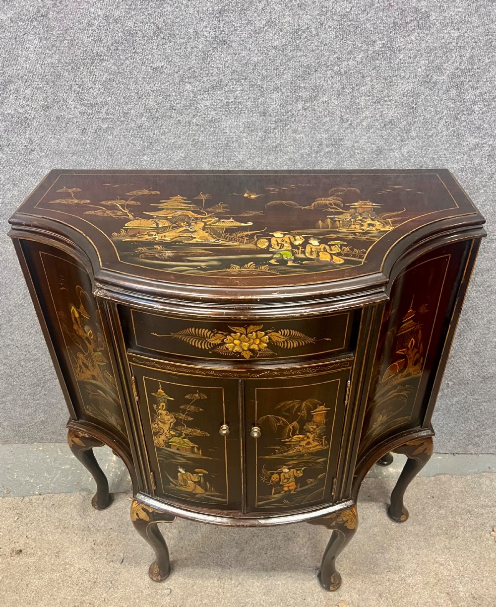 A dark lacquered bow-front Chinoiserie cabinet featuring gilded pictorial landscapes and cabriole legs.