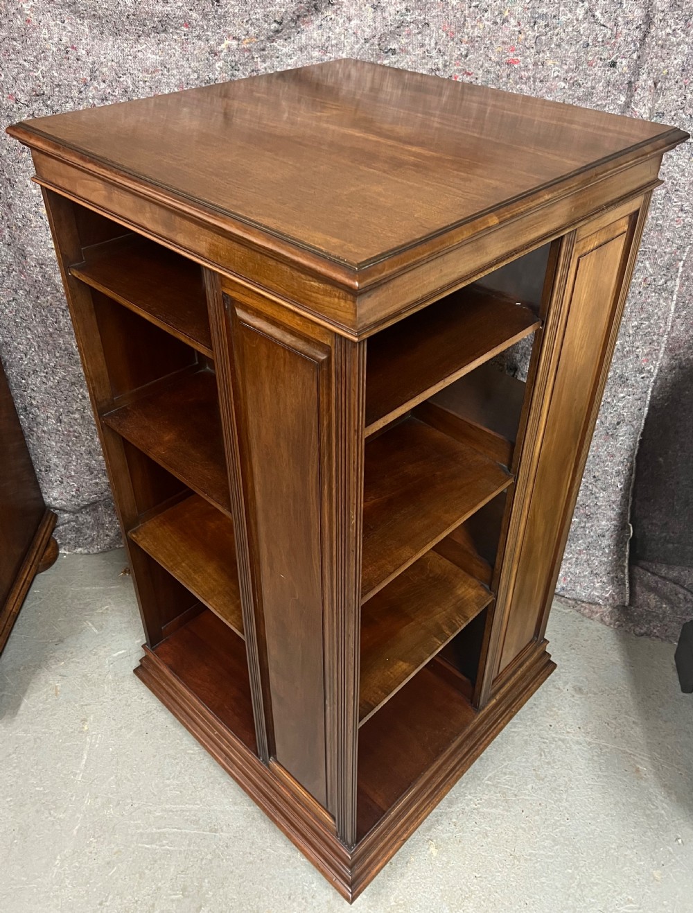 A four-sided warm brown wooden revolving bookcase featuring multiple tiers of open shelving, corner panel details, and a moulded top cornice. View III