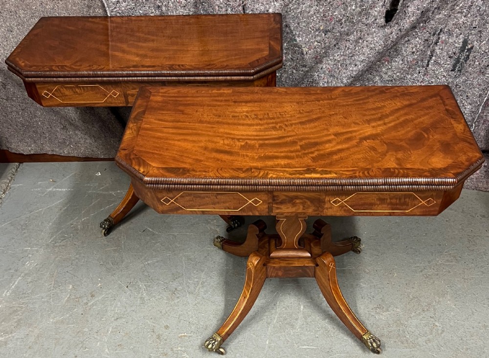 A pair of Regency flame mahogany fold-over card tables featuring boxwood inlay, reeded edges, and pedestal bases with splayed legs ending in brass paw castors.