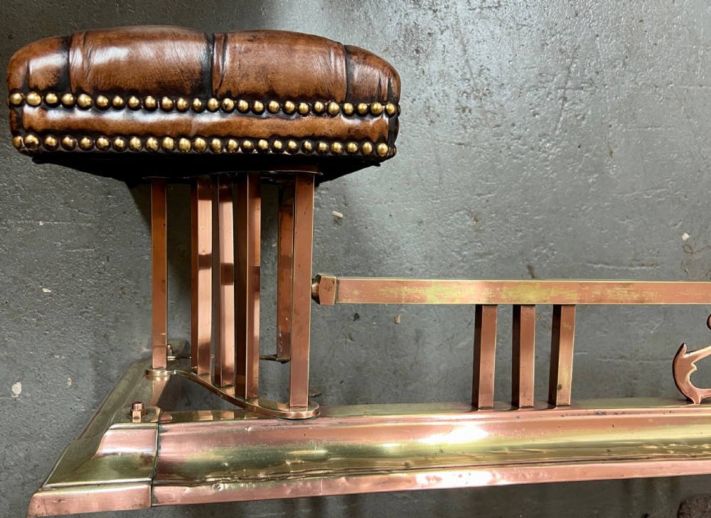 An antique brass and copper club fender featuring two button-tufted brown leather corner seats with brass stud detailing set on a low metal base. View V