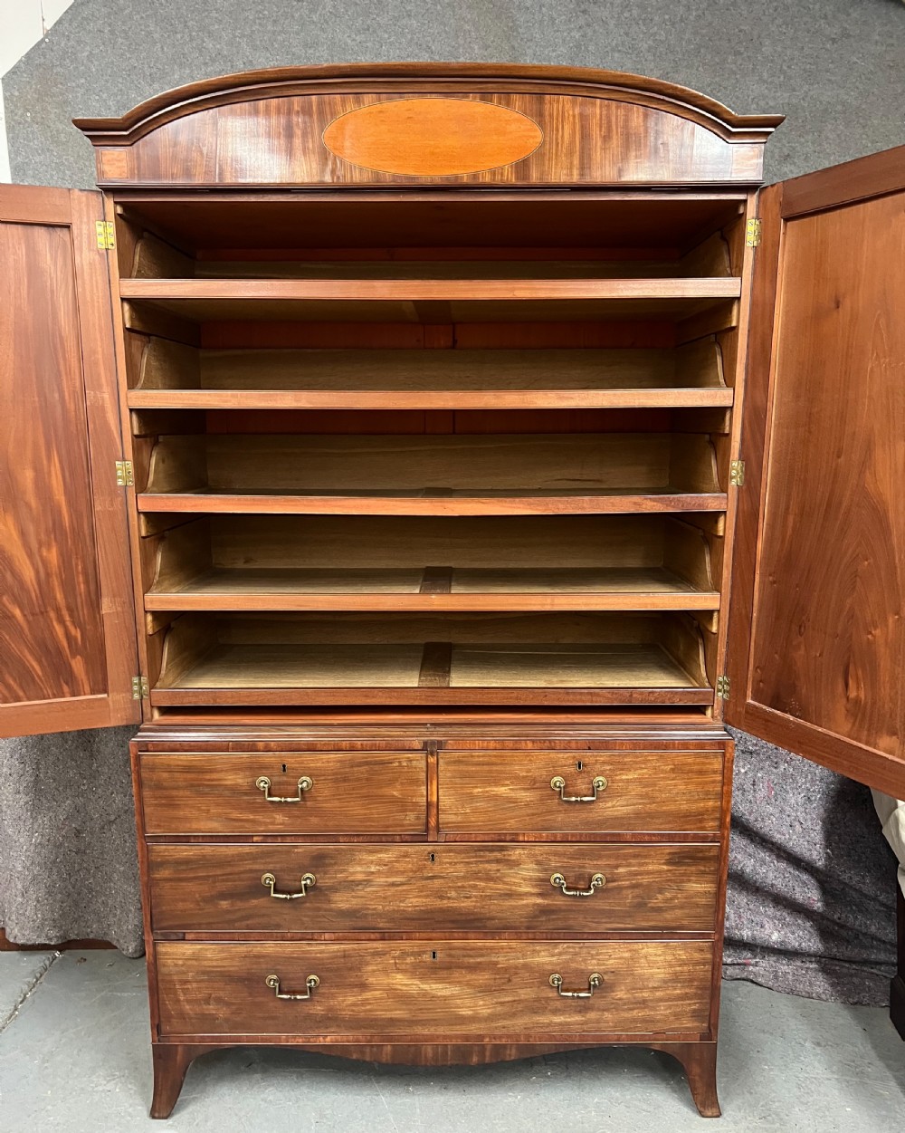 A George IV mahogany linen press featuring an arched pediment with oval inlay, double doors with oval flame mahogany panels, and a chest of drawers base. View IV
