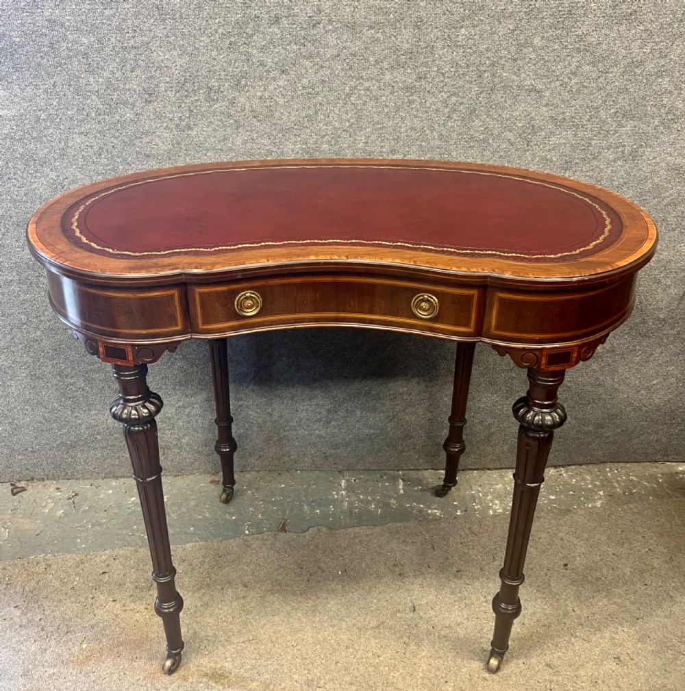 An Edwardian kidney-shaped mahogany writing table featuring a deep red leather insert with gilt tooling, satinwood string inlay, turned fluted legs, brass castors, and a central curved drawer.