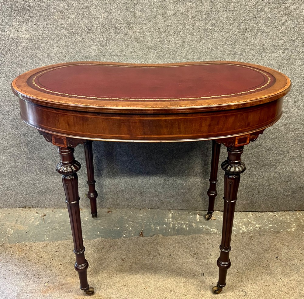 An Edwardian kidney-shaped mahogany writing table featuring a deep red leather insert with gilt tooling, satinwood string inlay, turned fluted legs, brass castors, and a central curved drawer. View IV