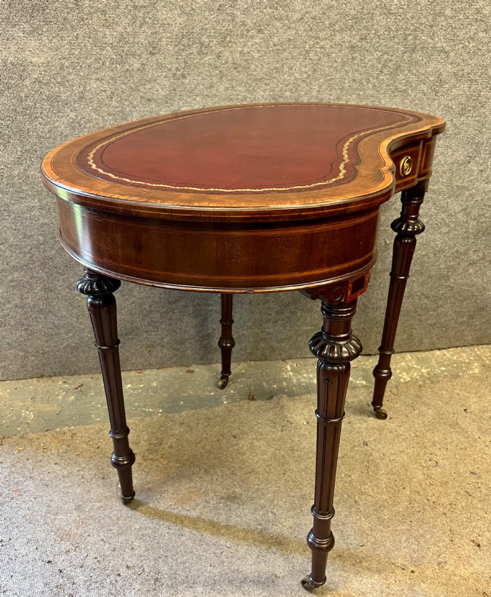 An Edwardian kidney-shaped mahogany writing table featuring a deep red leather insert with gilt tooling, satinwood string inlay, turned fluted legs, brass castors, and a central curved drawer. View V