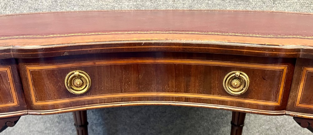 An Edwardian kidney-shaped mahogany writing table featuring a deep red leather insert with gilt tooling, satinwood string inlay, turned fluted legs, brass castors, and a central curved drawer. View IX
