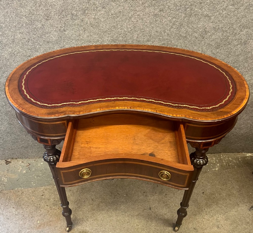 An Edwardian kidney-shaped mahogany writing table featuring a deep red leather insert with gilt tooling, satinwood string inlay, turned fluted legs, brass castors, and a central curved drawer. View X