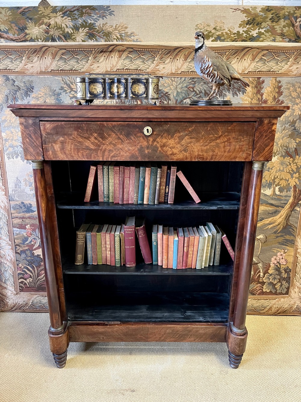 An antique French Empire open bookcase crafted from flame mahogany, featuring three open shelves, a top frieze drawer, and side column supports.
