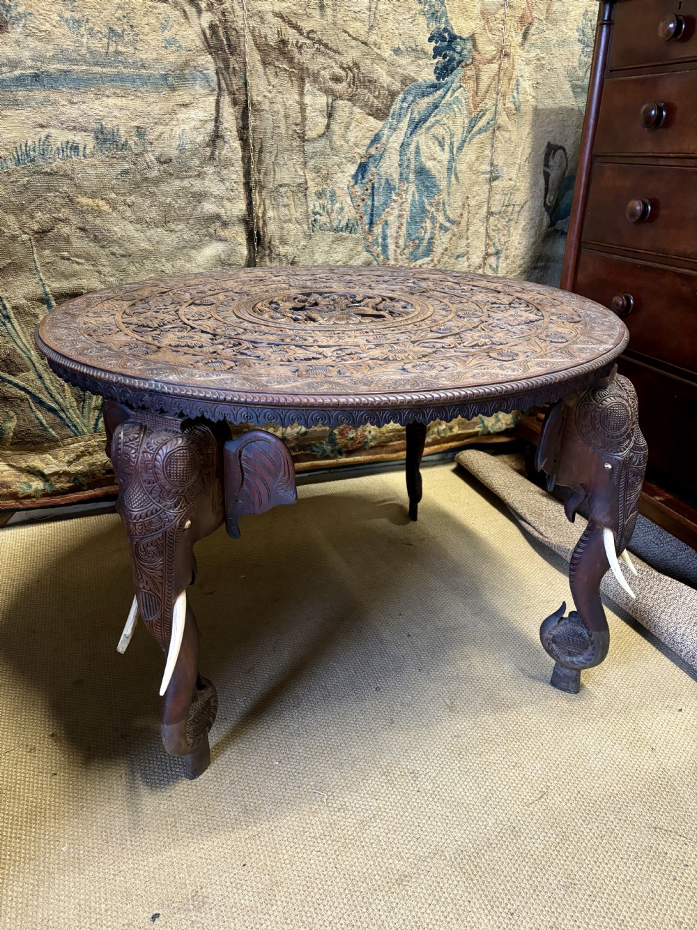 A large circular dark teak Anglo-Indian table featuring a deeply hand-carved floral top supported by three carved elephant legs with white tusks.