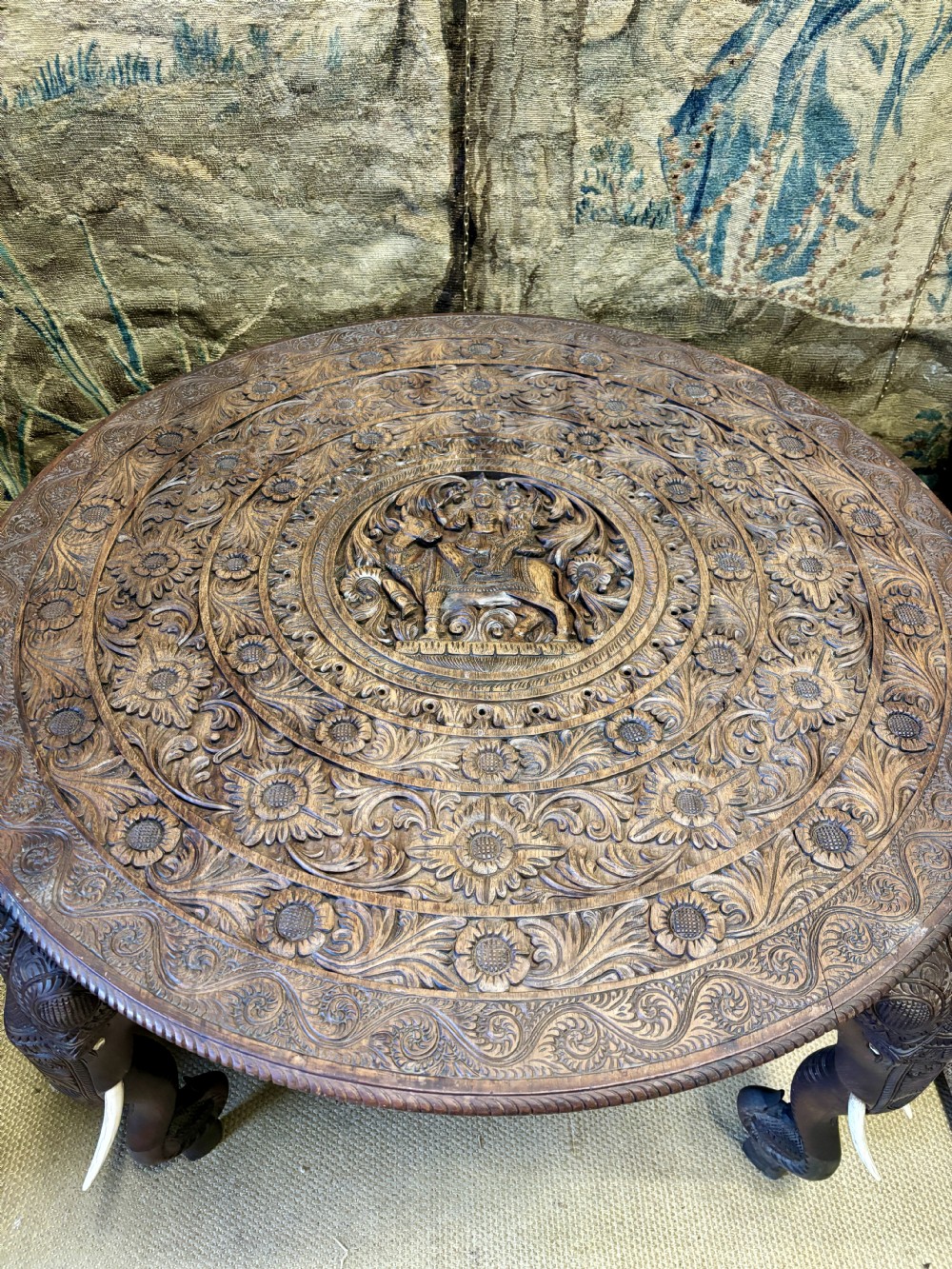 A large circular dark teak Anglo-Indian table featuring a deeply hand-carved floral top supported by three carved elephant legs with white tusks. View II