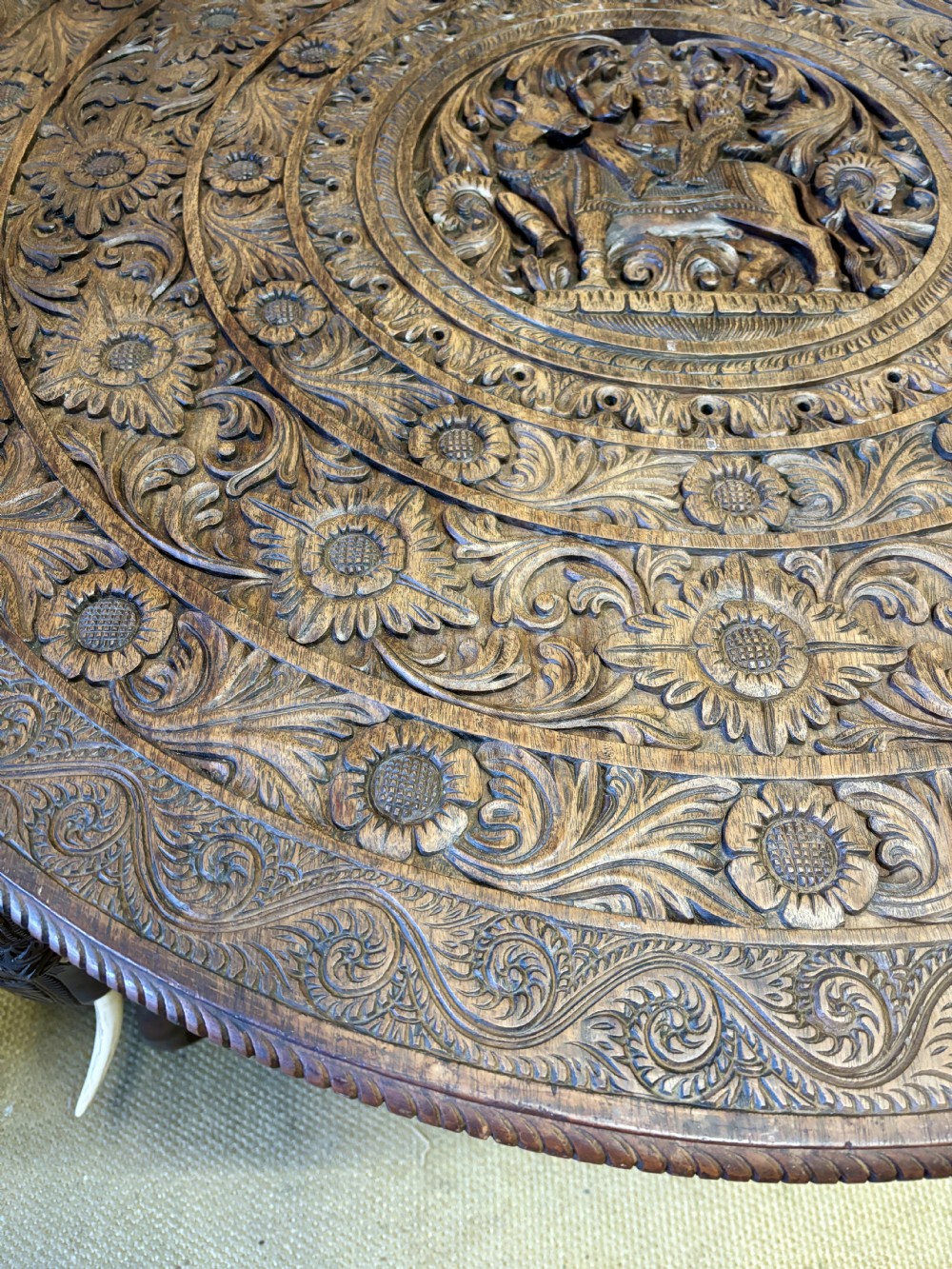 A large circular dark teak Anglo-Indian table featuring a deeply hand-carved floral top supported by three carved elephant legs with white tusks. View IV
