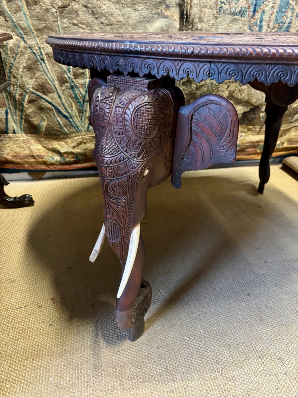 A large circular dark teak Anglo-Indian table featuring a deeply hand-carved floral top supported by three carved elephant legs with white tusks. View V