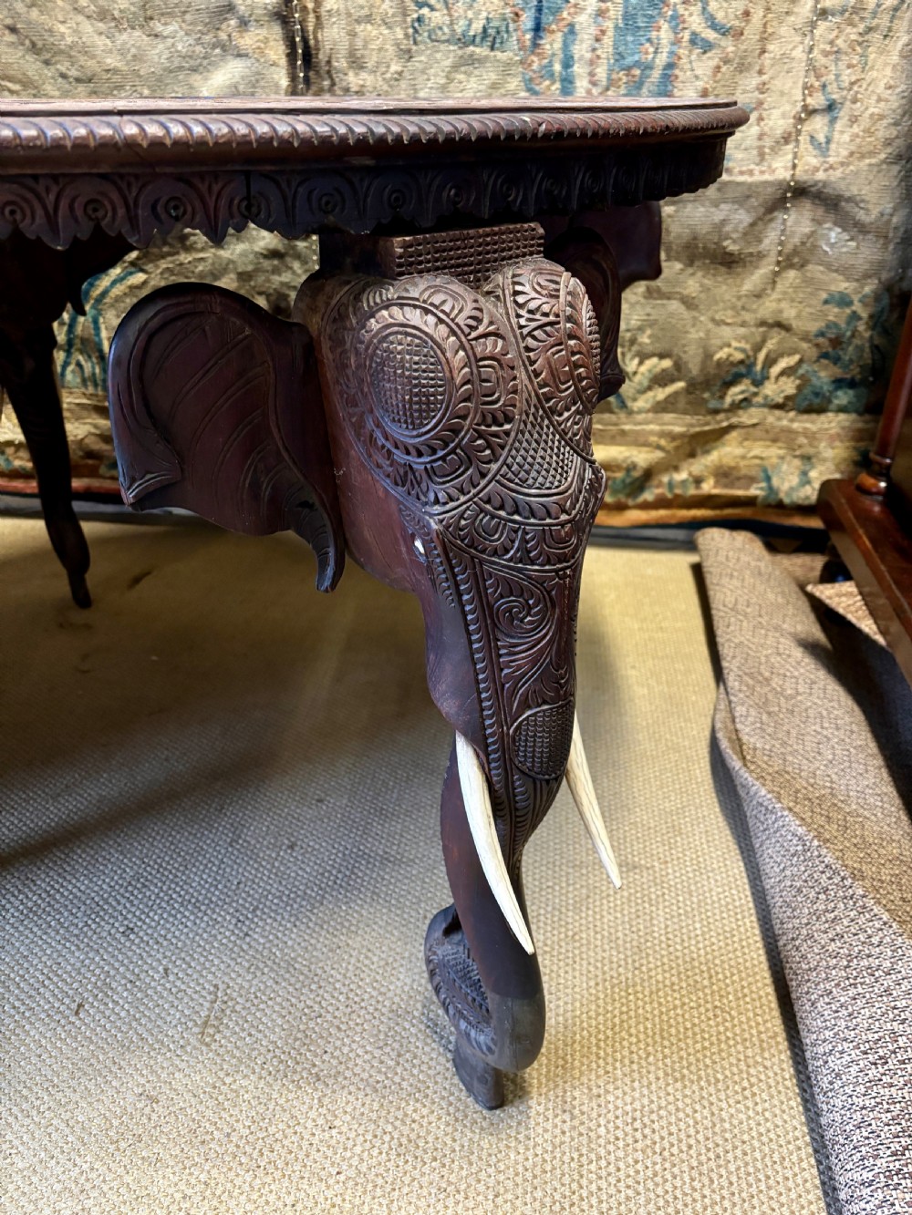 A large circular dark teak Anglo-Indian table featuring a deeply hand-carved floral top supported by three carved elephant legs with white tusks. View VI