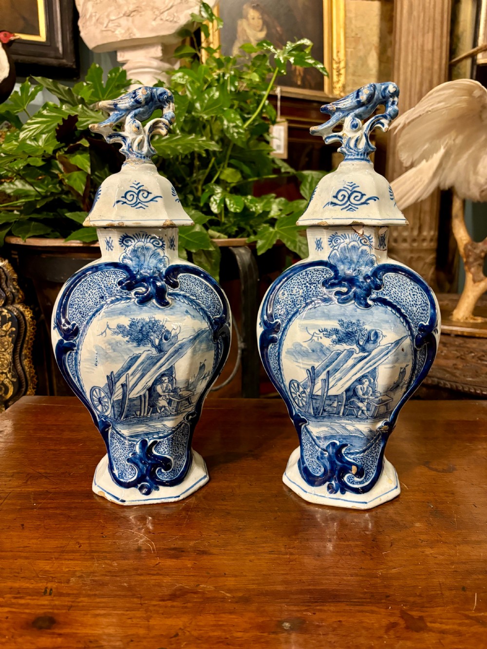 A pair of octagonal 18th-century Dutch Delftware lidded vases featuring blue hand-painted scenes within scrollwork cartouches and topped with bird finials.