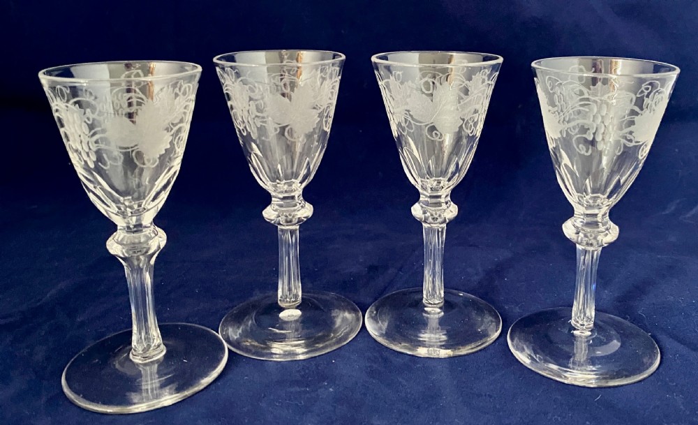 A set of four clear Victorian glass cordial glasses featuring acid-etched grape and vine motifs around the bowls and faceted stems.