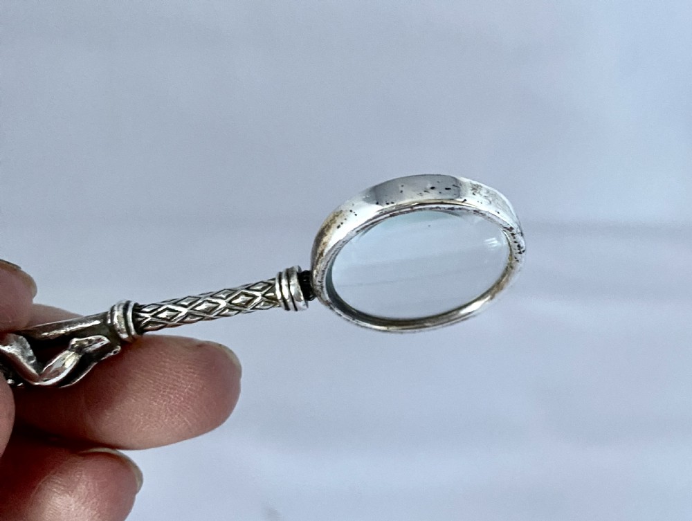 An antique silver-mounted magnifying glass featuring a sculpted cherub handle with a diamond-textured stem above a circular glass lens. View V