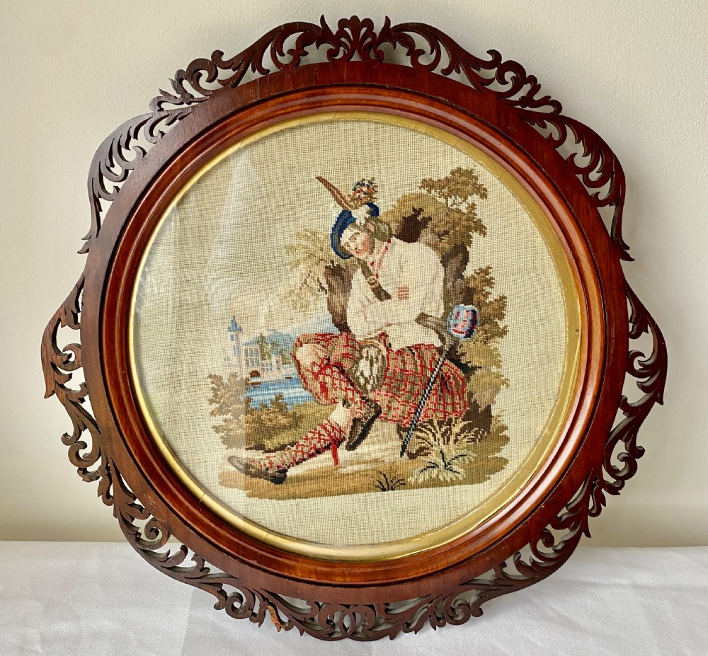 A circular Victorian woolwork picture depicting a seated Scottish Highlander in a kilt, housed in a pierced carved wooden frame.