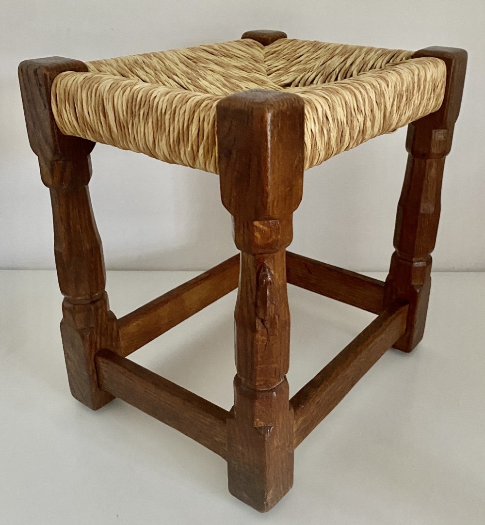 An English Arts and Crafts solid oak stool featuring a woven rush seat, turned faceted legs connected by rectangular stretchers, and a small carved mouse motif on one leg.