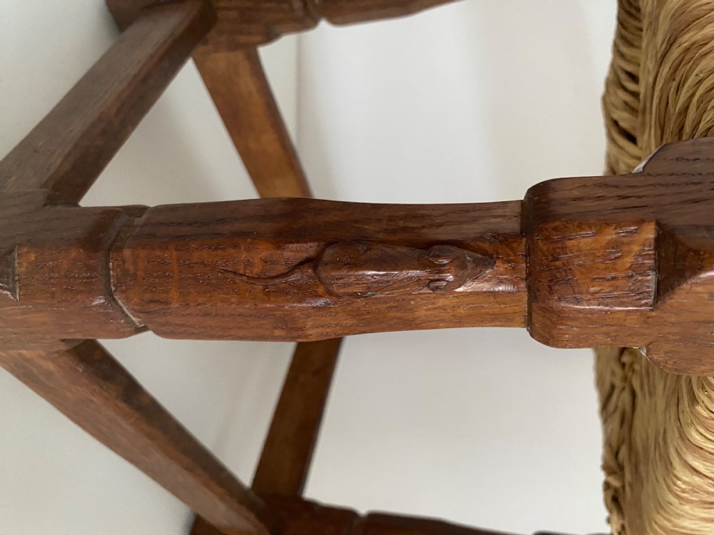 An English Arts and Crafts solid oak stool featuring a woven rush seat, turned faceted legs connected by rectangular stretchers, and a small carved mouse motif on one leg. View III