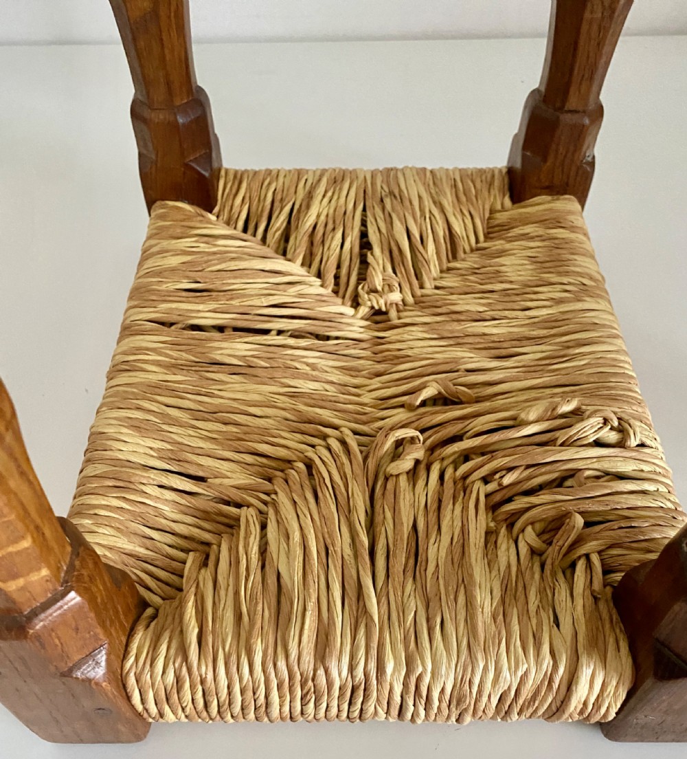 An English Arts and Crafts solid oak stool featuring a woven rush seat, turned faceted legs connected by rectangular stretchers, and a small carved mouse motif on one leg. View IV