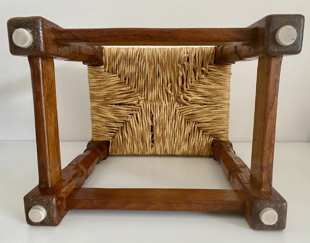 An English Arts and Crafts solid oak stool featuring a woven rush seat, turned faceted legs connected by rectangular stretchers, and a small carved mouse motif on one leg. View VI