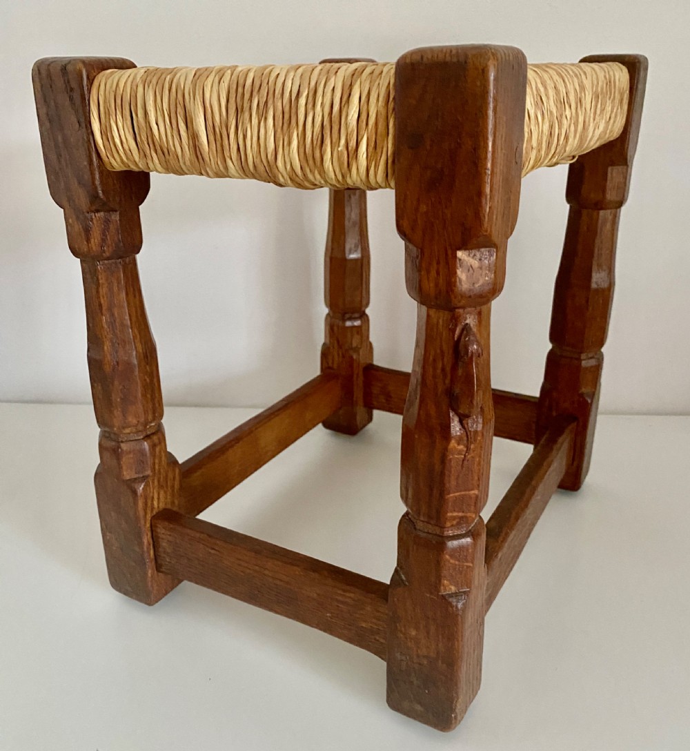 An English Arts and Crafts solid oak stool featuring a woven rush seat, turned faceted legs connected by rectangular stretchers, and a small carved mouse motif on one leg. View VII