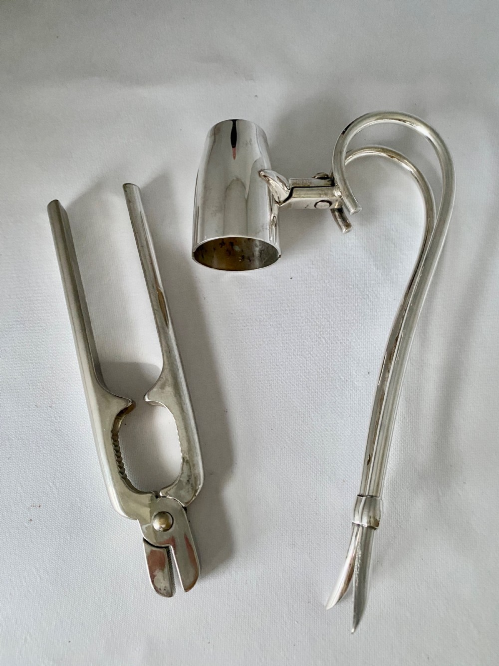 A silver-plated Art Deco champagne bottle pouring handle and matching champagne pliers set on a white fabric surface.