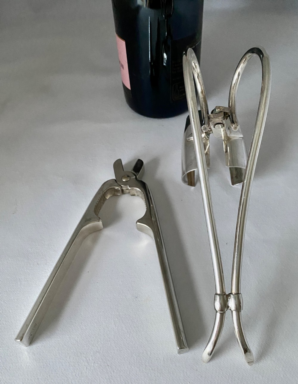 A silver-plated Art Deco champagne bottle pouring handle and matching champagne pliers set on a white fabric surface. View V