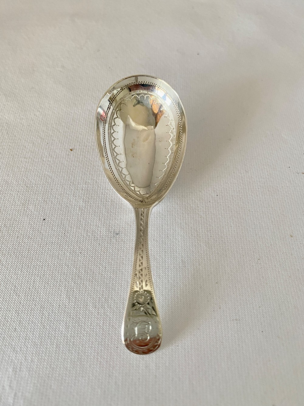 An antique sterling silver caddy spoon featuring an oval bowl with bright-cut engraved borders and a monogrammed handle.