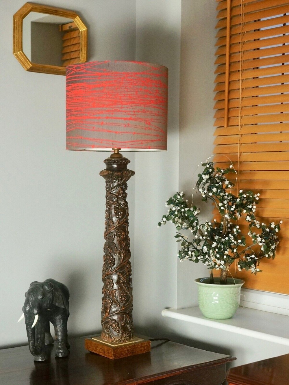 A tall hand-carved dark wooden table lamp base featuring relief fruiting vine motifs, fitted with a grey and red patterned drum shade.