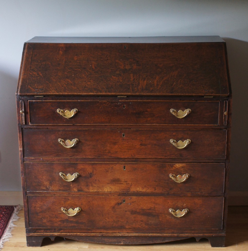 An 18th-century dark oak bureau featuring a slanted fall-front writing flap above four graduated drawers with brass handles.